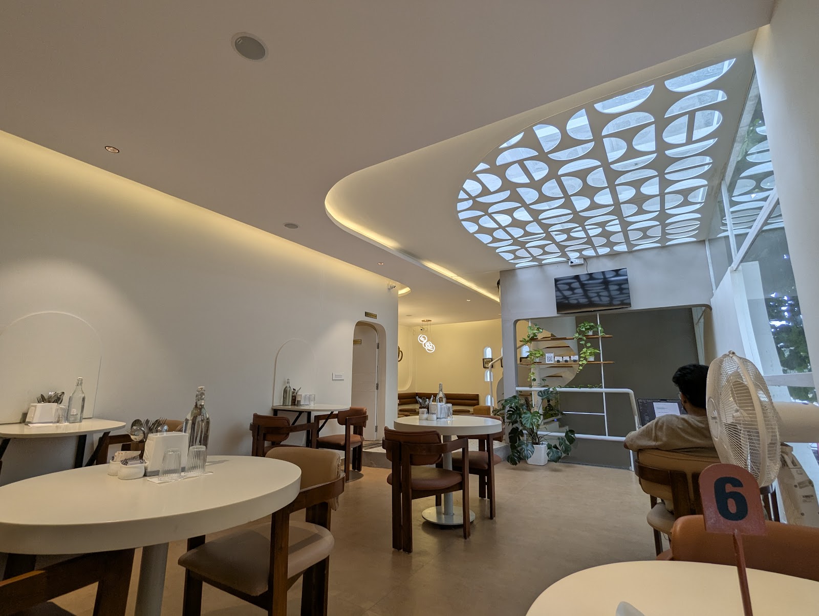 The main dining room in daylight: white walls, round tables with cane-backed chairs, and a perforated lattice panel let into the ceiling above the stair.