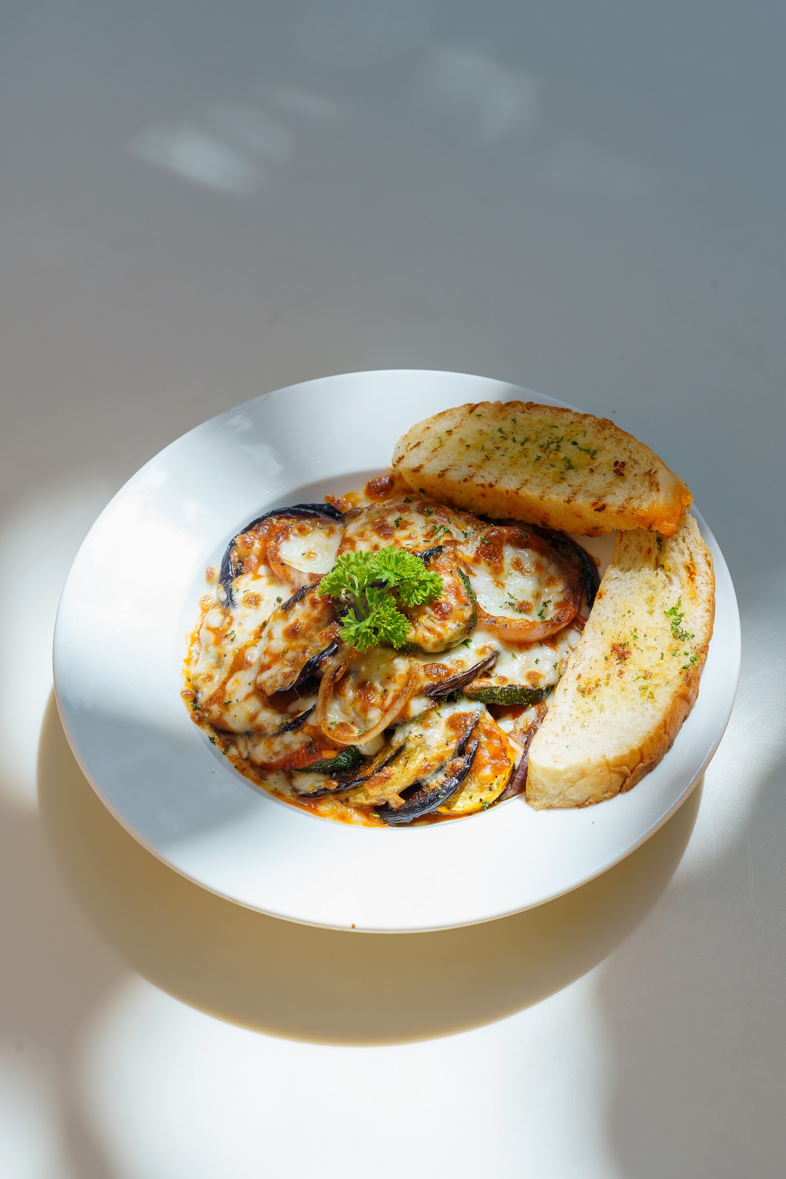 A white plate of grilled aubergine slices scattered with herbs, served with wedges of toasted bread, lit by hard afternoon sun.