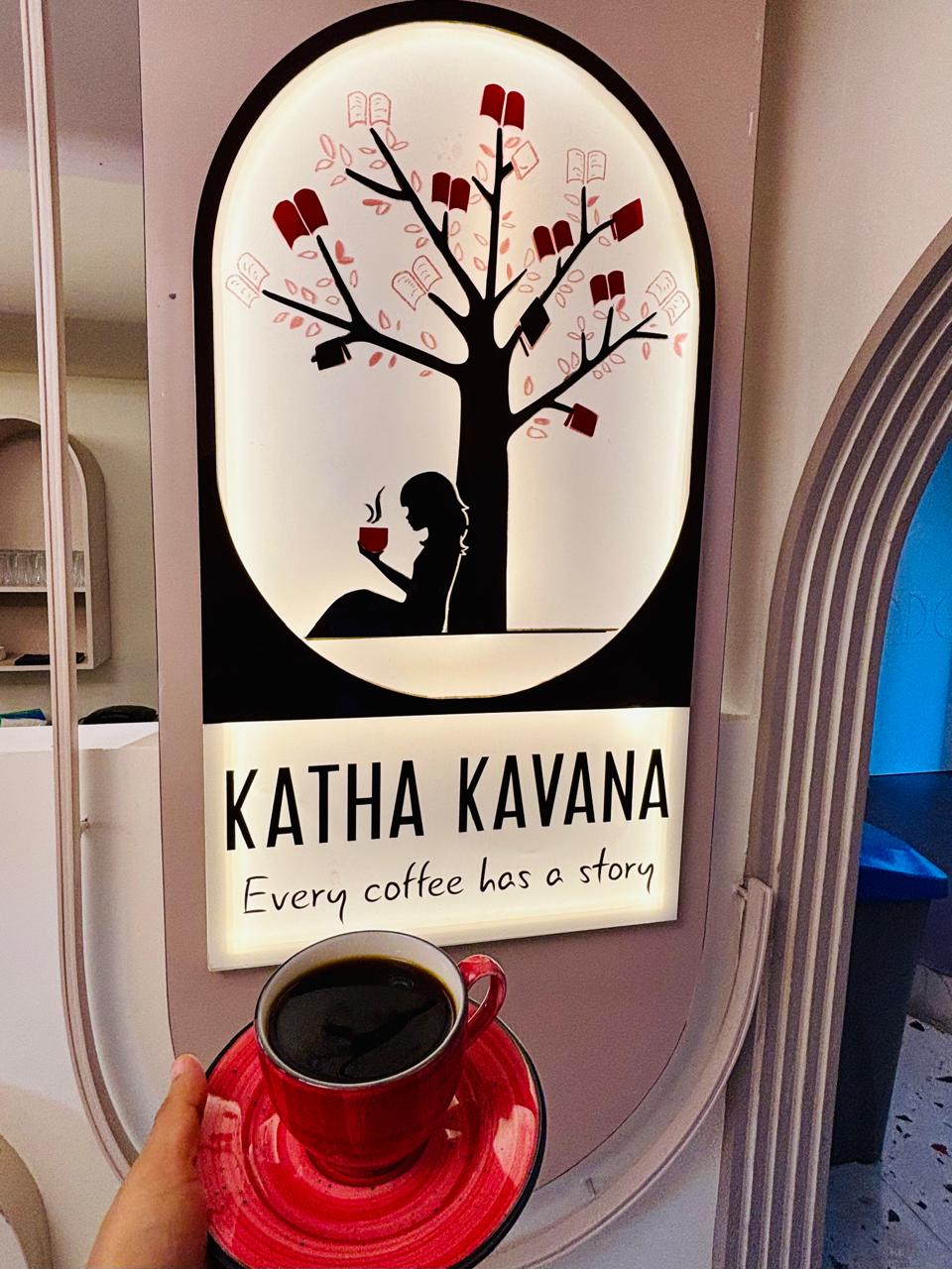 A hand holds a small red cup of black coffee in front of the painted house sign: a tree hung with books, a reader sitting under it, and the words Katha Kavana, every coffee has a story.