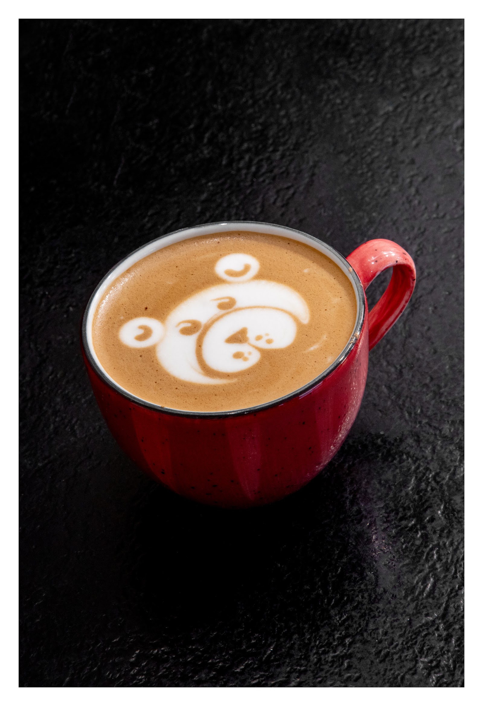 A red cup of cappuccino on a black surface, its foam drawn into a bear's face.