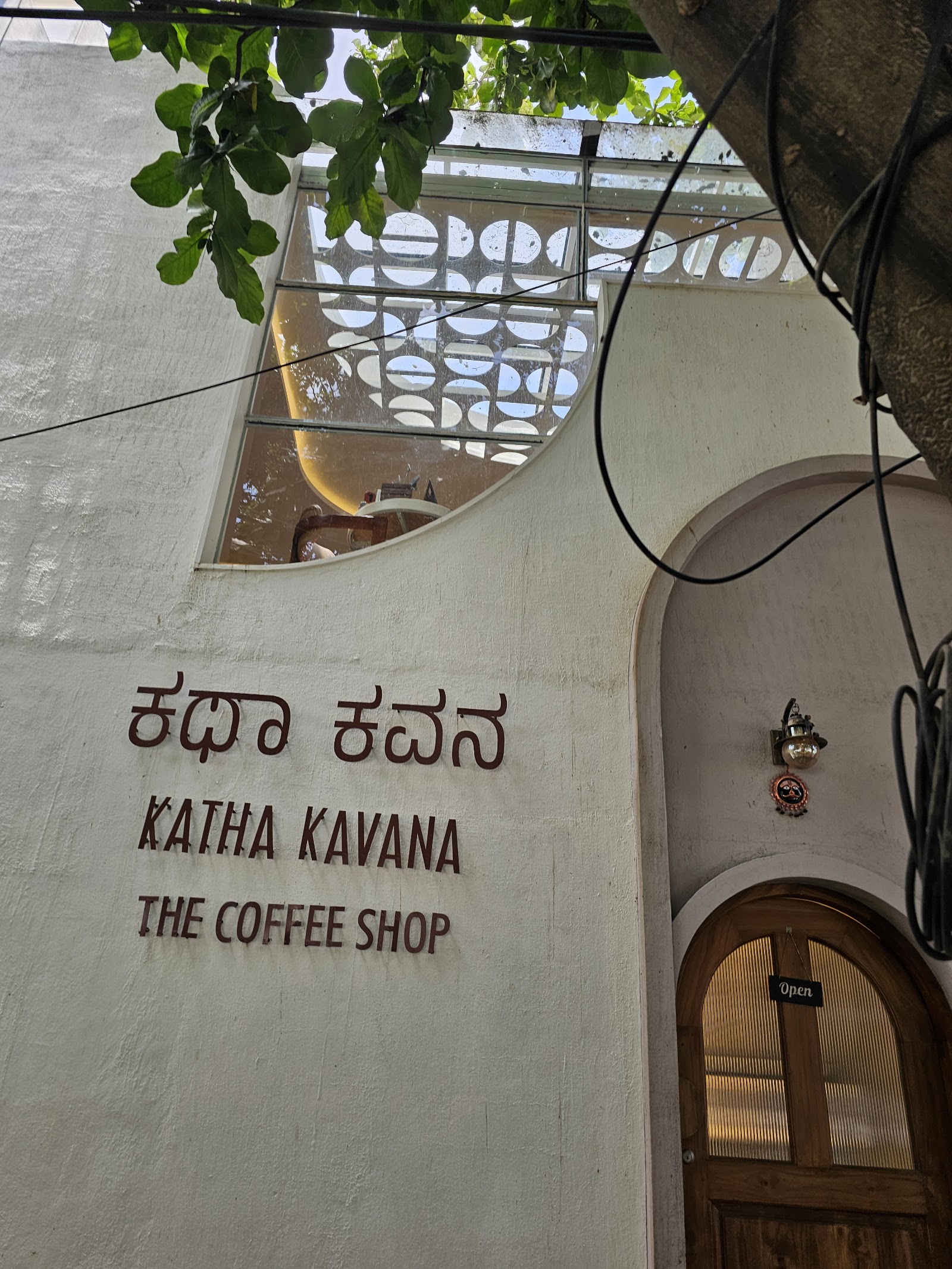 The outside wall in daylight: the name painted in Kannada as ಕಥಾ ಕವನ above the roman Katha Kavana and the words the coffee shop, with leaves overhanging an arched window.