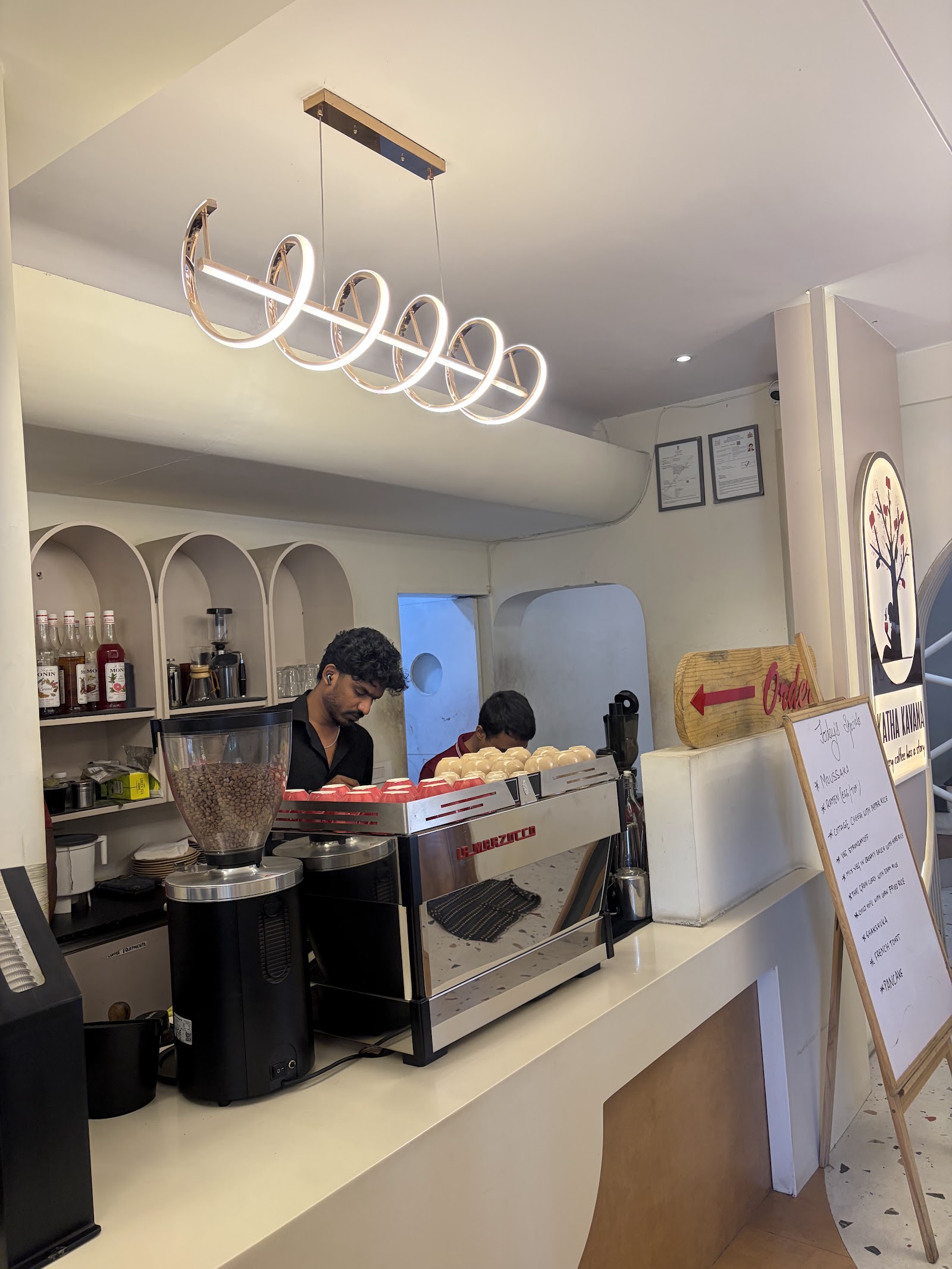 The service counter, a barista working behind it, brewing equipment and grinders ranged along the white top under a coiled pendant light.