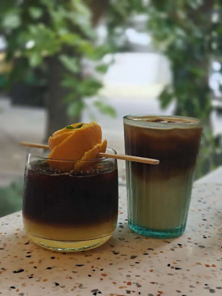 Two tall iced drinks on a terrazzo table under leaves — one dark, garnished with a dried orange slice, one pale green.