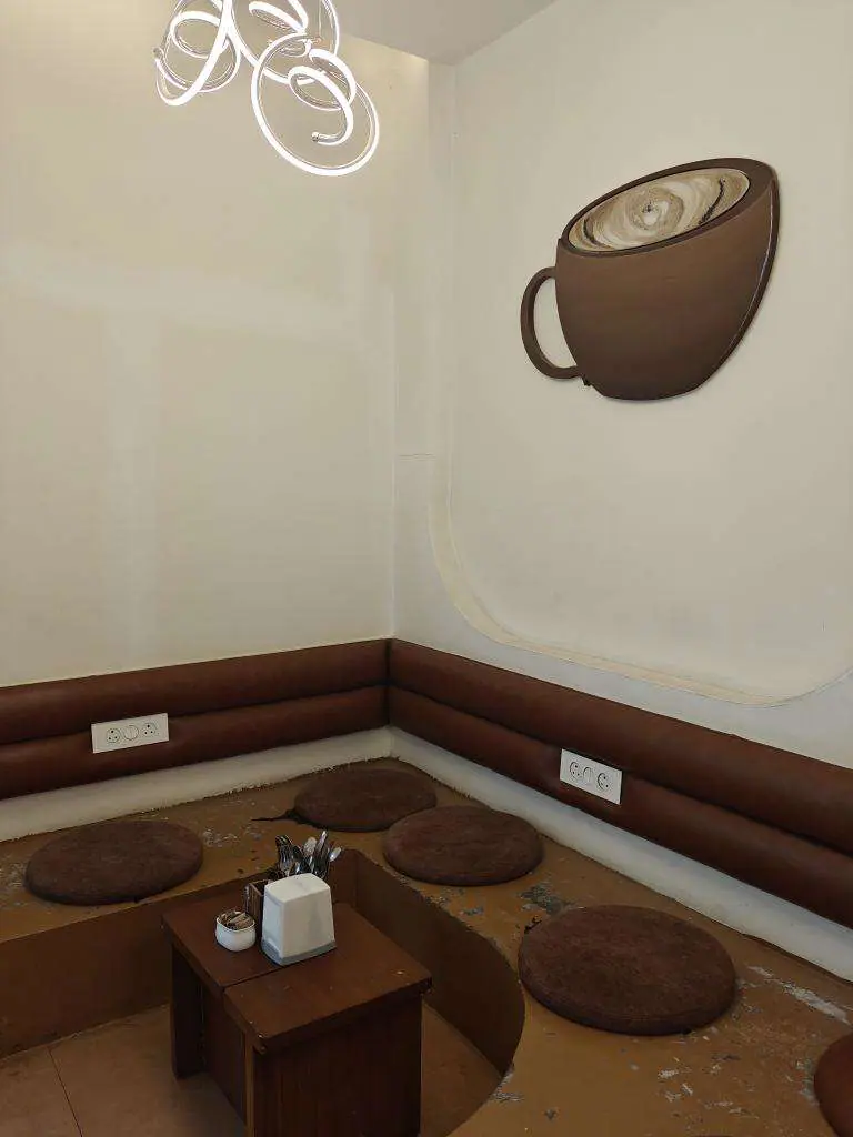 A quiet corner: a bench banquette, low round tables, and a coffee cup drawn on the wall in a single line with steam rising from it.