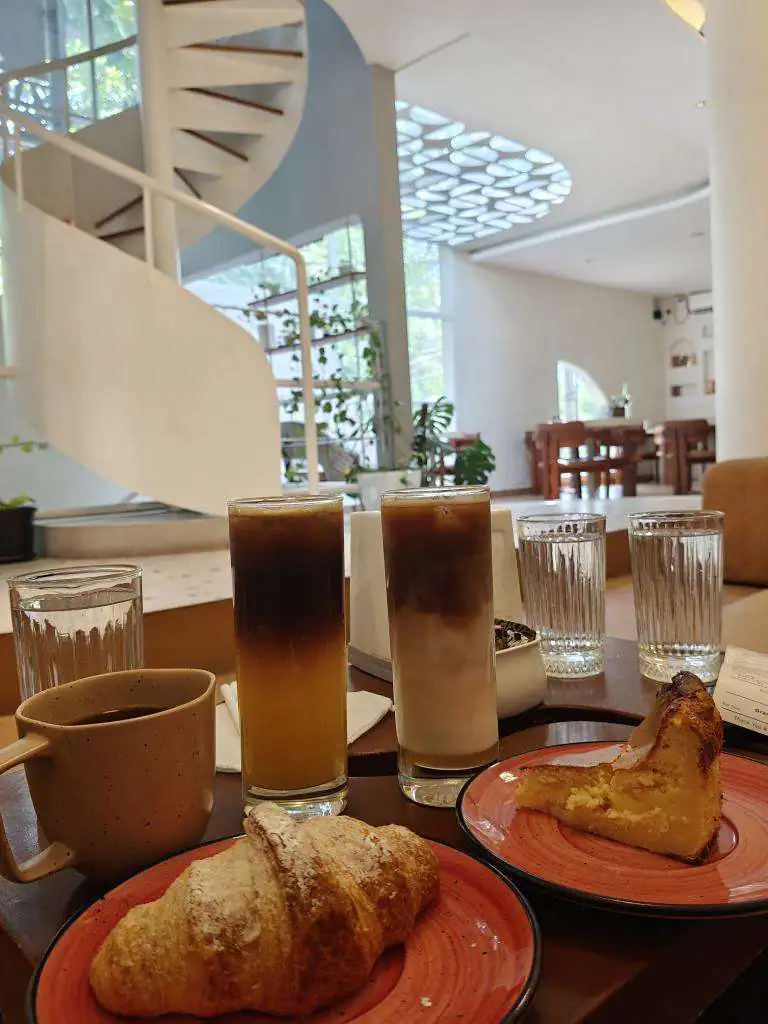 A table of iced coffees with a bun and a pastry on a red plate, the spiral staircase rising behind.