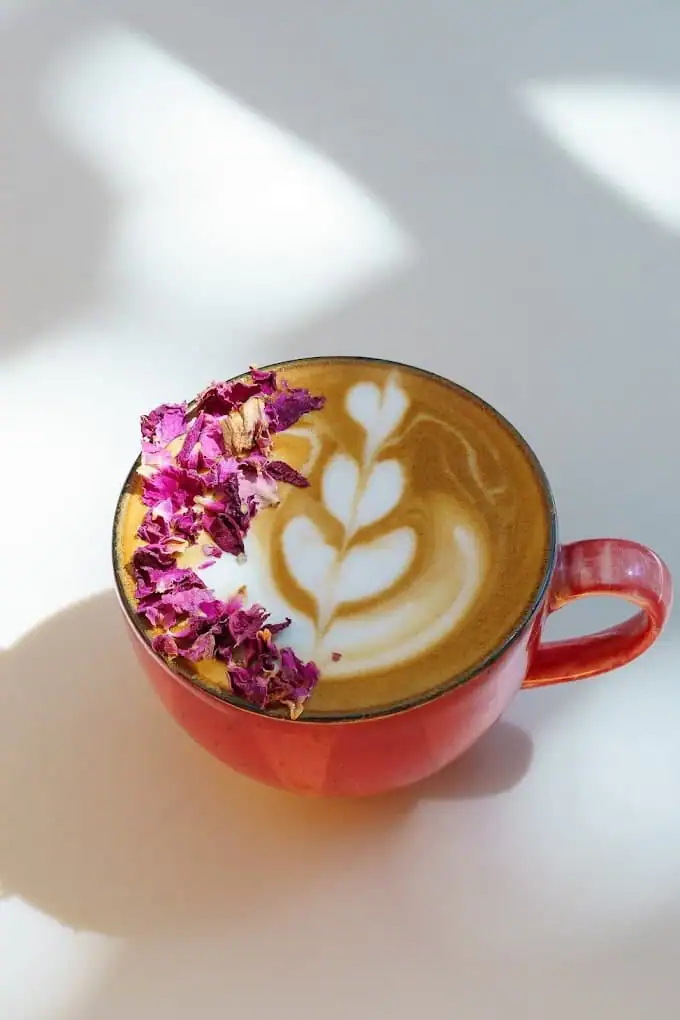 A latte in a red cup with a leaf poured into the foam, dried flowers laid on the saucer, on a white table in dappled light.