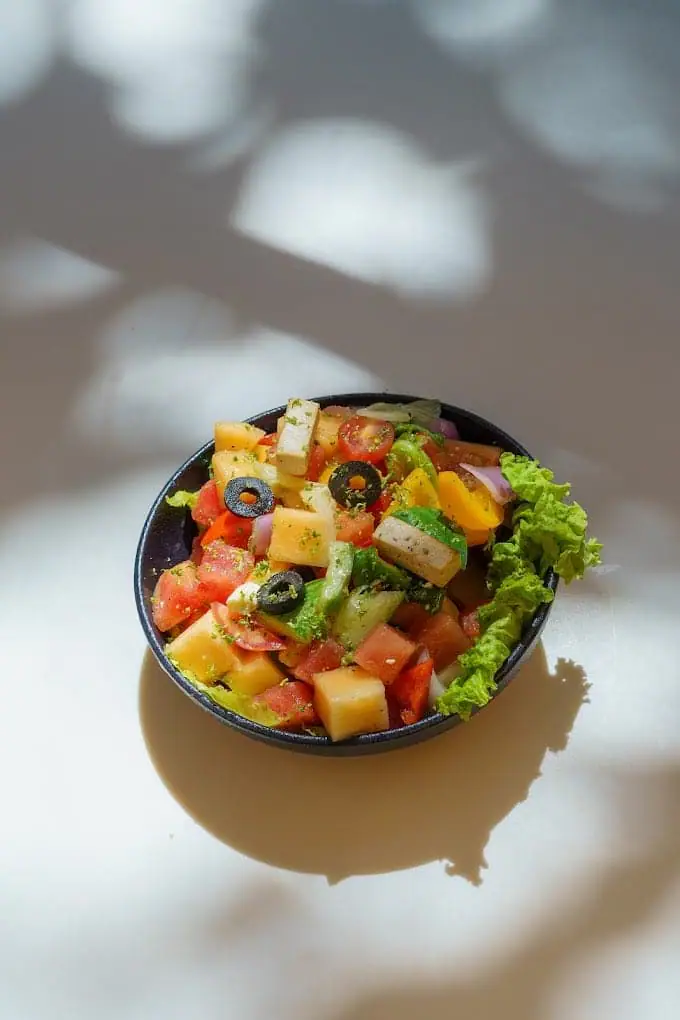 A dark bowl of chopped salad — tomato, cucumber, black olives and lettuce — on a white table.