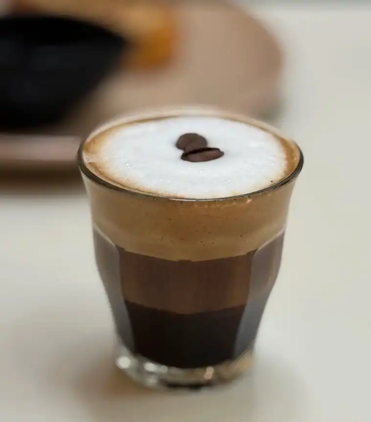 A layered iced coffee in a straight glass under a thick cap of milk foam, a single coffee bean set on top.