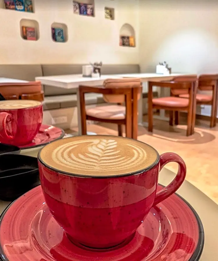 A large red cup and saucer of cappuccino with a rosetta poured into the foam, dining tables and wooden chairs out of focus behind it.