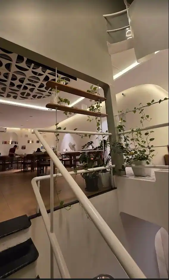 The view from the mezzanine — white railing, trailing plants, the staircase, and seating on the floor below under the lattice ceiling.